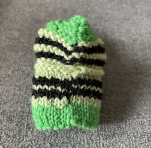Back view of a green and black striped knitted baby bootie, showing how the stripes don't meet up, on a gray cloth surface.